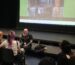 Phil sitting on stage, with images of tattoos in background and learners seated in theatre.