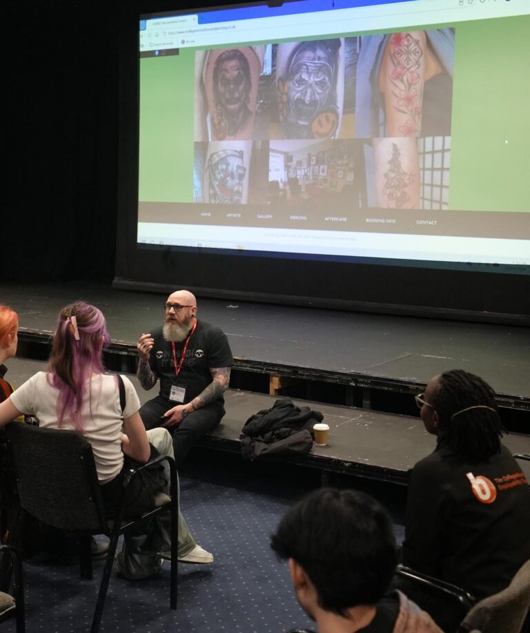 Phil sitting on stage, with images of tattoos in background and learners seated in theatre.